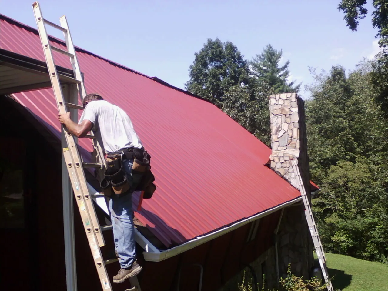 Roofer on a ladder beside a red metal roof and stone chimney — Soffit & Fascia Repair by Roofing Brothers in Spotsylvania